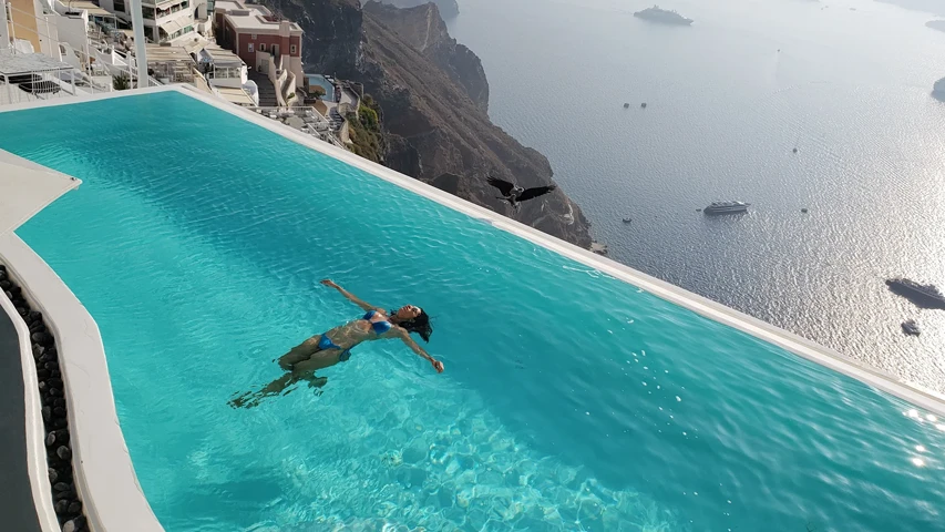 Santorini'nin nefes kesici kaldera manzarasına bakan lüks bir otel olan Cosmopolitan Suites'in sonsuzluk havuzunda sırtüstü rahatça yüzen bir kadın, havuzun kenarından denize açılan geniş ufuk ve gökyüzünde süzülen bir kuş görülüyor.