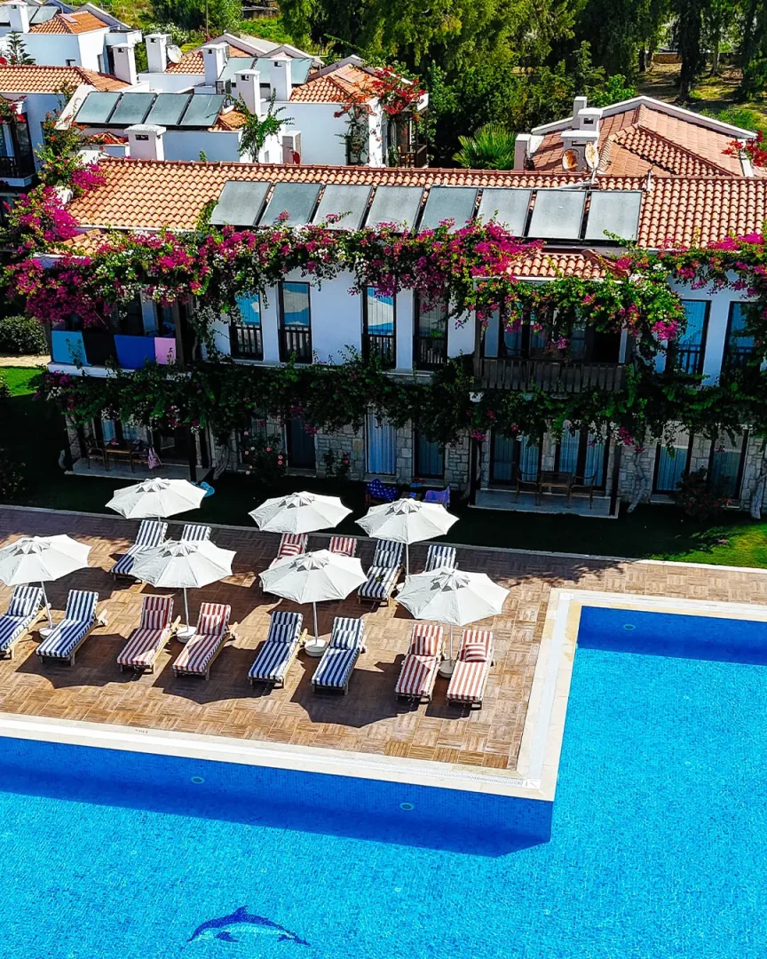 Datça Doris Hotel'in yemyeşil ağaçlarla çevrili tesisinde, binaları saran pembe begonviller, kiremit çatılardaki güneş panelleri, yunus mozaikli pırıl pırıl mavi bir yüzme havuzu ve etrafındaki beyaz şemsiyeler ile çizgili şezlongların huzurlu bir kuşbakışı görünümü.