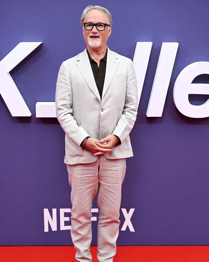 Yönetmen David Fincher, gri saçları, gözlükleri ve beyaz sakalıyla, açık bej rengi keten takım elbisesi ve koyu renk polo yaka tişörtü içinde, elleri kenetli bir şekilde kırmızı halıda poz veriyor; mor arka planda kısmen KİLLER ve Netflix logoları görülüyor.