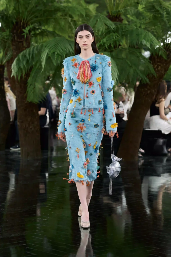 Model, palmiye ağaçlarıyla çevrili yansıtıcı bir zeminde yürürken, Jonathan Anderson'ın Dior Sonbahar-Kış 2026 Haute Couture koleksiyonundan, Ahmedabad'ın zanaat mirası ve Lynda Benglis'in heykelsi yaklaşımlarından ilham alan, canlı renklerde çiçek aplikeleri, püsküller ve boncuklarla süslü açık mavi tüvit ceket ve etek takımını, gümüş rengi stilize bir tavus kuşu çantasıyla sergiliyor.