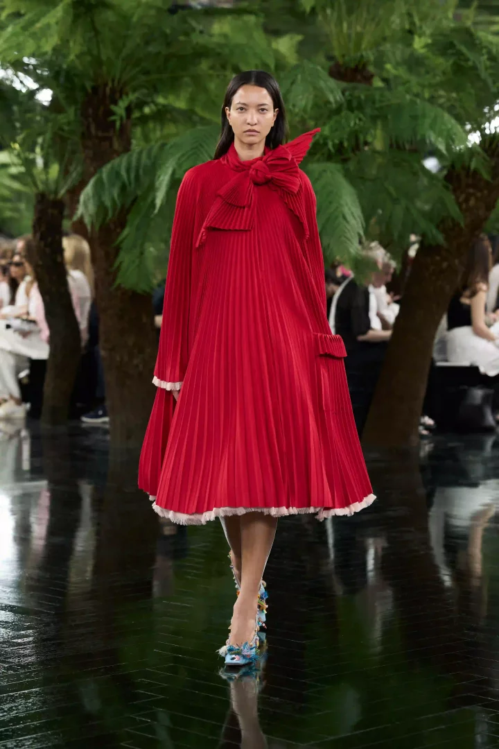 Palmiye ağaçları arasında, Dior Sonbahar-Kış 2026 Haute Couture koleksiyonundan heykelsi duruşlu, hacimli kırmızı plili elbisesi ve çok renkli çiçek aplikeli ayakkabılarıyla yürüyen bir model, Jonathan Anderson'ın kumaşı heykele dönüştüren zanaat yaklaşımını Lynda Benglis'in sanatsal pratiği ve Ahmedabad'ın zanaat mirasıyla buluşturuyor.