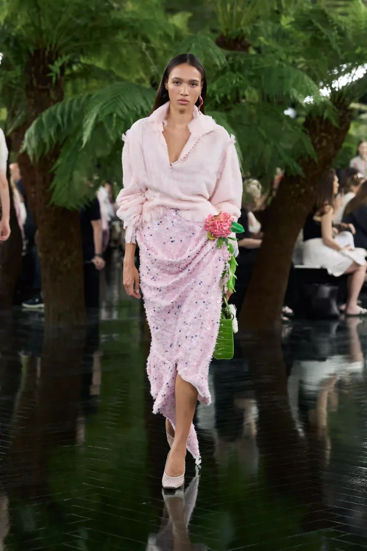 Jonathan Anderson'ın Dior Sonbahar-Kış 2026 Haute Couture koleksiyonundan bir model, yemyeşil bitki örtüsüyle çevrili parlak podyumda, pembe dokulu bluzu ve asimetrik kesimli, pul işlemeli eteğinin yanına iliştirilmiş büyük pembe çiçek aplikesi ve yeşil çantasıyla, Lynda Benglis'in heykelsi drapajlarından ilham alan couture zanaatini sergiliyor.