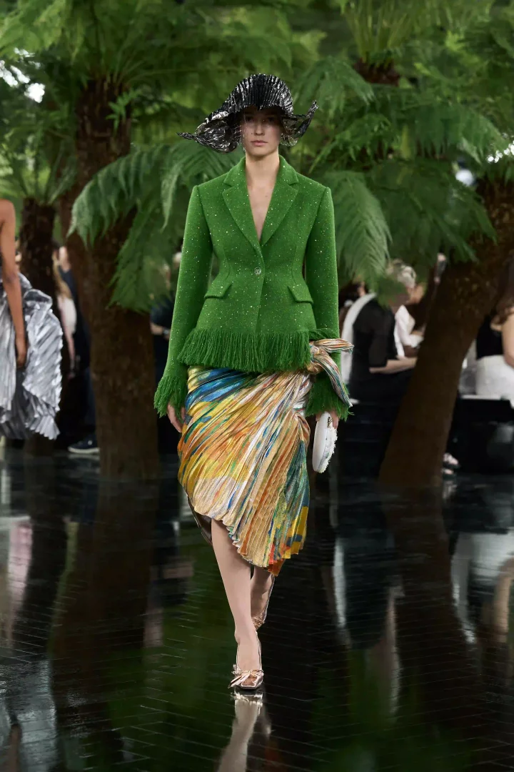 Jonathan Anderson'ın Dior Sonbahar-Kış 2026 Haute Couture koleksiyonundan bir model, Lynda Benglis'in heykelsi formlarını anımsatan, parıltılı yeşil püsküllü ceketi ve Ahmedabad'ın canlı renklerini yansıtan, pilili ve drapeli etekle podyumda yürürken, zanaat mirasının deneysel bir ifadesini sergiliyor.