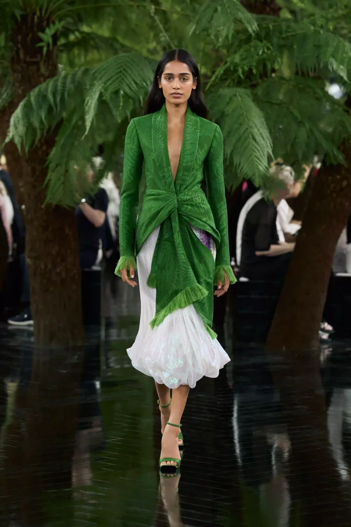 Dior Sonbahar-Kış 2026 Haute Couture koleksiyonundan bir model, Lynda Benglis'in heykelsi formlarından ilham alan, derin V yakalı yeşil, düğümlü üst kısmı ve parlak beyaz, hacimli eteğiyle doğa temasını yansıtan bir kıyafet içinde podyumda yürüyor.