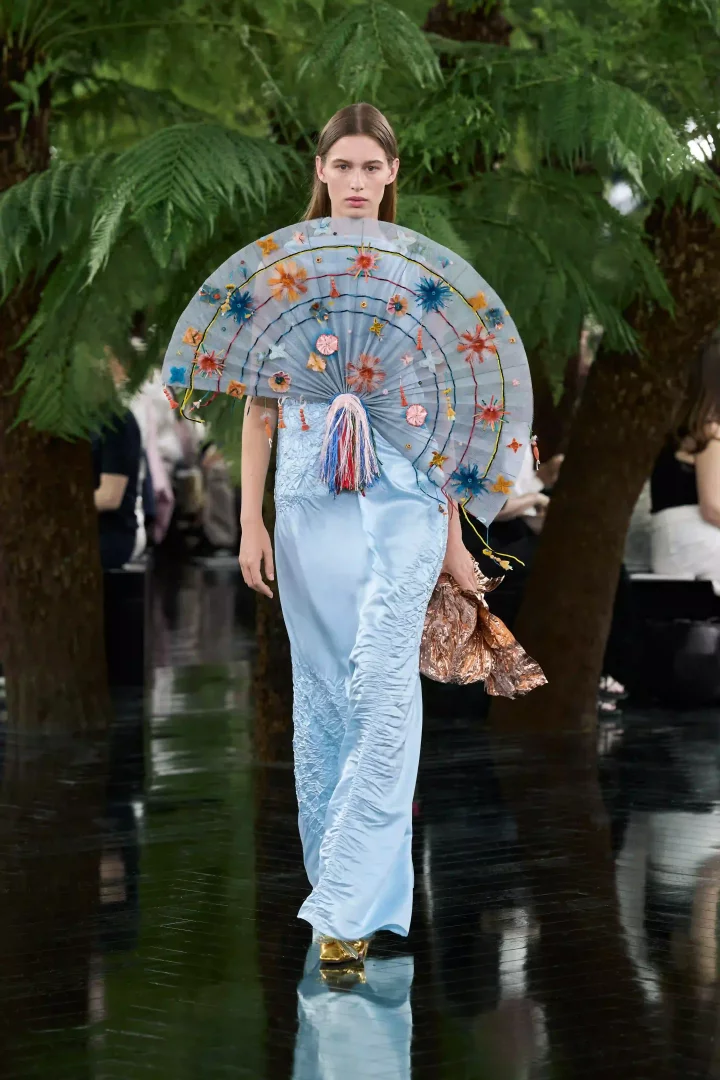 Dior Sonbahar-Kış 2026 Haute Couture koleksiyonundan bir manken, Rodin Müzesi'nin bahçelerini anımsatan yeşil bitkilerle çevrili, yansıtıcı podyumda yürüyor; Jonathan Anderson'ın sanatsal dokunuşunu yansıtan, uçuk mavi pileli elbisesi ve zengin boncuk işlemeli, Japon yelpazesini andıran heykelsi göğüs aksesuarıyla dikkat çekiyor.
