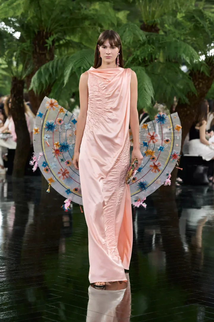 Dior Sonbahar-Kış 2026 Haute Couture defilesinde, yeşil ağaçlarla çevrili Rodin Müzesi bahçelerindeki yansıtıcı podyumda yürüyen bir model, açık pembe, drape ve büzgü detaylı uzun bir elbise giyiyor; arkasında Hint zanaatından ilham alan, işlemeli ve püsküllü yelpaze şeklinde göz alıcı bir aksesuar taşıyor.