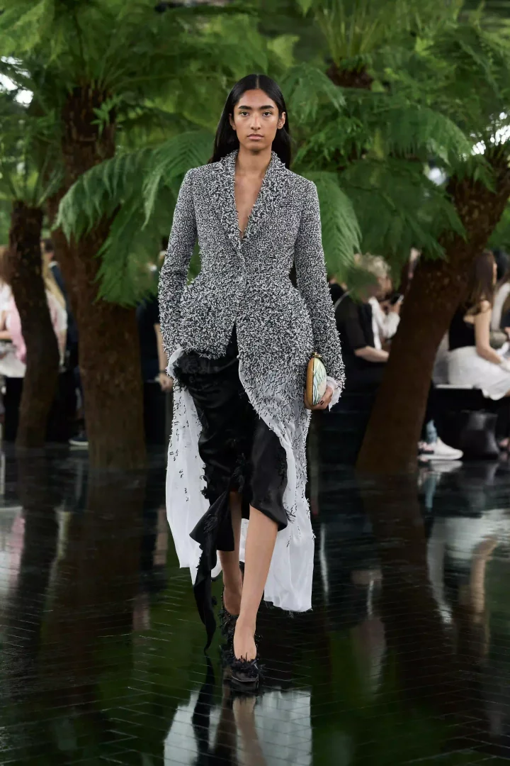 Dior Sonbahar-Kış 2026 haute couture defilesinde bir model, Rodin Müzesi'nin yemyeşil bahçeleri temalı yansıtıcı podyumda, Jonathan Anderson imzalı, dokulu gri-beyaz peplum bir ceket ile akışkan siyah ve şeffaf beyaz katmanlı etek kombinasyonunu, işlemeli bir minik clutch ile tamamlayarak zarifçe ilerliyor.