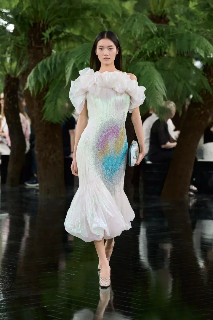 Paris Haute Couture Moda Haftası'nda, Dior Sonbahar-Kış 2026 koleksiyonundan Jonathan Anderson imzalı, omuzları volanlı, fildişi rengi ışıltılı, balık kuyruğu formundaki elbisesiyle bir model, ortasındaki rengarenk pul işlemelerle Rodin Müzesi'nin bahçelerindeki yansıtıcı podyumda göz kamaştırıyor.