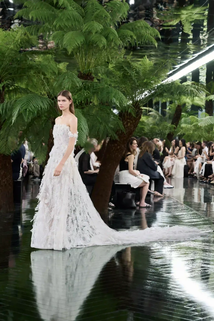 Dior Sonbahar-Kış 2026 Haute Couture defilesinde, Rodin Müzesi'nin yemyeşil bahçelerini yansıtan parlak zeminde, tüylü ve zarif detaylara sahip, omuzları açık beyaz bir elbise giyen model podyumda yürüyor, koleksiyonun heykelsi ve doğa temalı couture anlayışını vurguluyor.