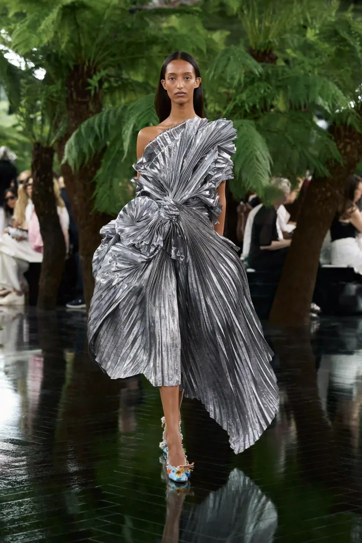 Rodin Müzesi'nin yemyeşil bahçelerinde, yansıtıcı zeminde yürüyen bir model, Jonathan Anderson'ın Lynda Benglis'ten ilham alan Dior Sonbahar-Kış 2026 couture koleksiyonundan, metalik yüzeyleri ve heykelsi pileleme teknikleriyle adeta yaşayan bir heykele dönüşen, tek omzu açık, gümüş bir elbise sunuyor.