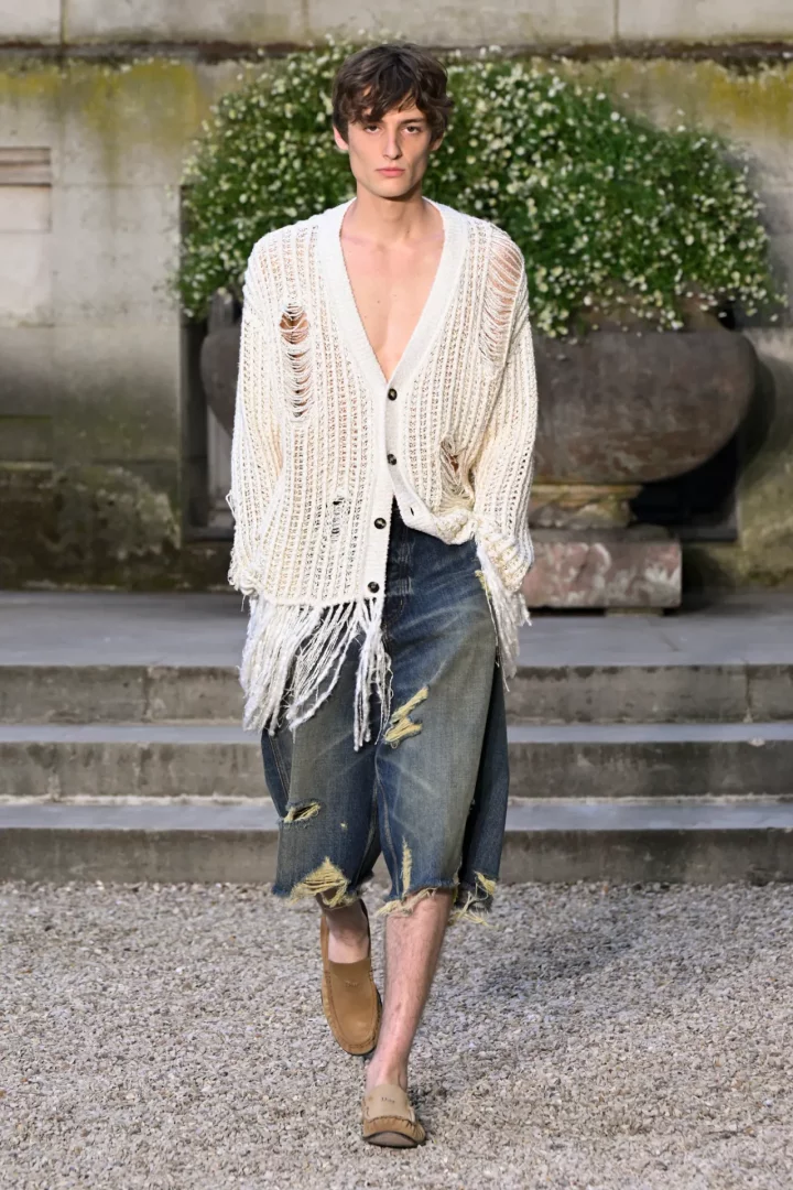 Paris Moda Haftası kapsamında Dior İlkbahar/Yaz 2027 erkek koleksiyonundan bir görünümü sergileyen, salaş ve yırtık detaylı, beyaz örgü hırka ile paçaları püsküllü ve yıpranmış kot şort giymiş genç bir erkek model, taş merdivenlerin önündeki çakıllı yolda yürüyor.