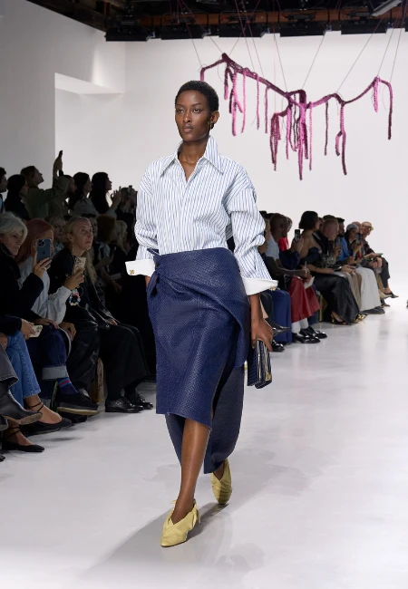 Podyumda yürüyen model, 2026 yaz etek trendlerinden biri olan diz hizası dokulu lacivert anvelop kalem eteğini, beyaz çizgili oversize gömlek ve sarı düz babetlerle kombinleyerek modern ve şık bir görünüm sunuyor.