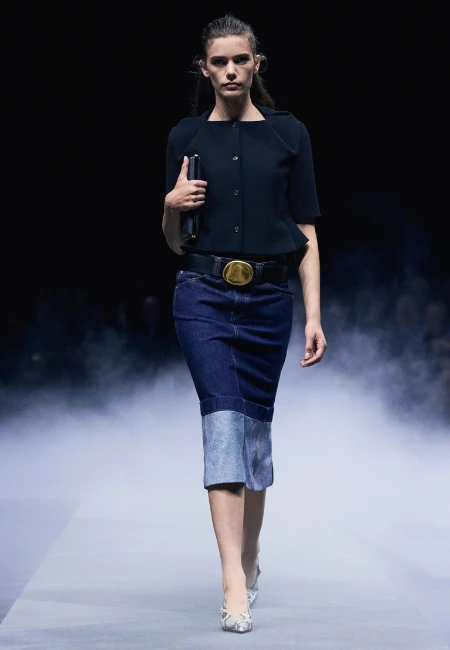 Podyumda yürüyen bir model, gold tokalı siyah bir kemerle tamamladığı koyu lacivert ve açık mavi iki renkli denim midi kalem etek ve kısa kollu siyah bluzuyla 2026 yaz etek trendlerini sergiliyor.