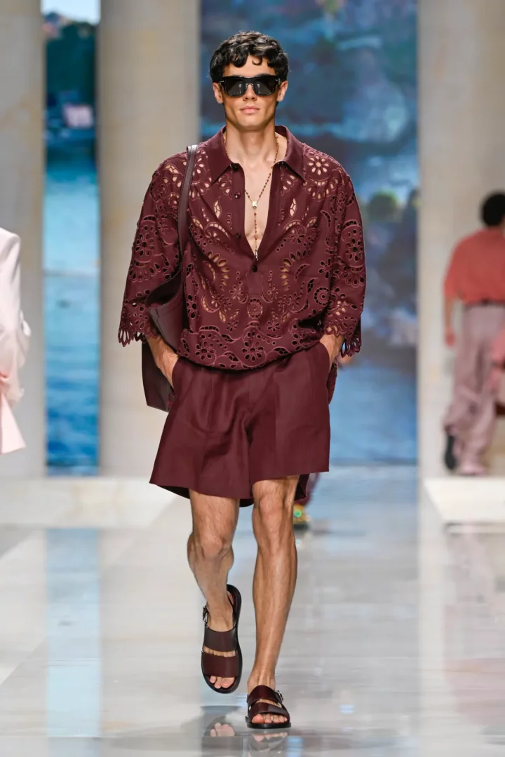 Dolce & Gabbana İlkbahar/Yaz 2027 erkek defilesinde podyumda yürüyen model, lazer kesim işlemeli bordo renkli kısa kollu gömleği, uyumlu şortu, sandaletleri, güneş gözlükleri ve uzun boncuklu kolyesiyle elegan bir yaz stili sergiliyor, elleri ceplerinde kendine güvenle ilerliyor.