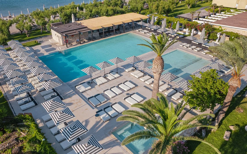 Samos'taki Doryssa Seaside Resort'un havuz alanını gösteren bu kuşbakışı fotoğrafta, açık mavi sulara sahip büyük havuzlar, şık siyah beyaz çizgili şemsiyelerle sıralanmış konforlu şezlonglar ve yemyeşil palmiye ağaçları denizin kıyısında keyifli bir atmosfer sunuyor.