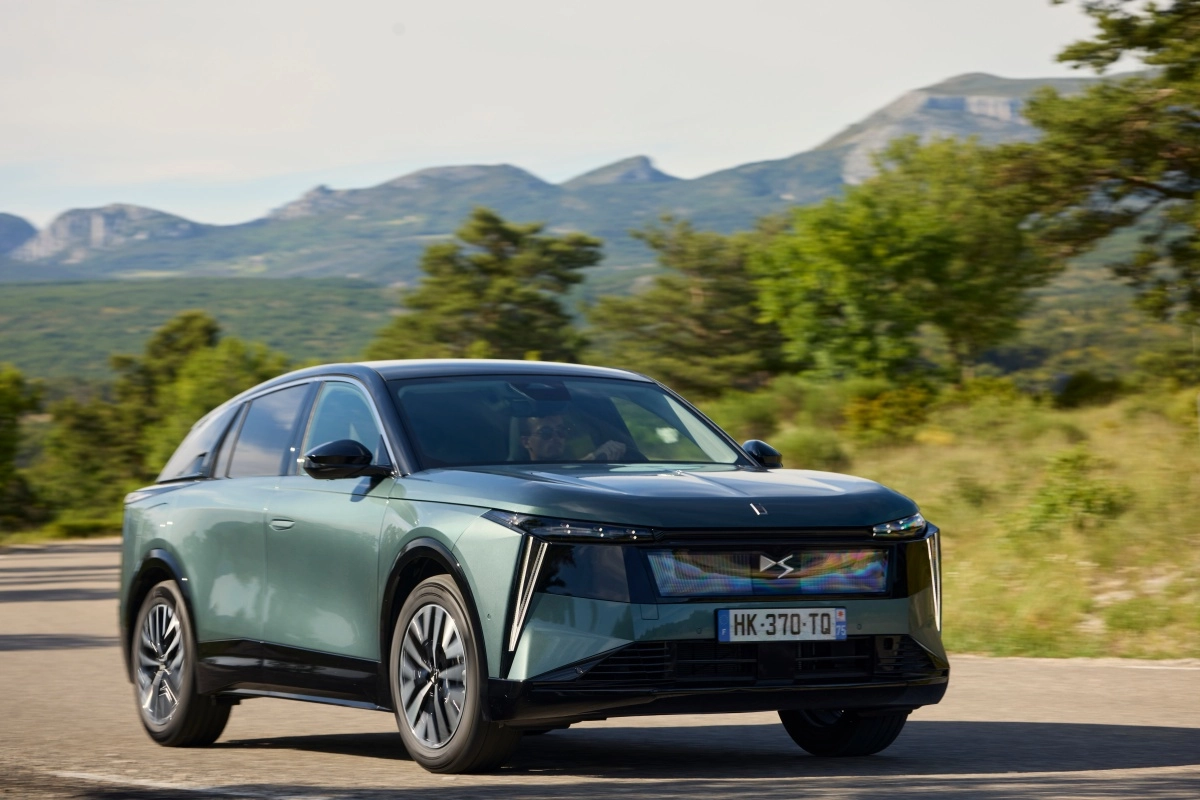 DS N7 lüks elektrikli premium SUV, metalik yeşilimsi gri rengi, belirgin dikey farları ve modern ön tasarımıyla, dağlık bir arka plana sahip ağaçlık yolda ileri doğru hareket ederken dikkat çekici bir şekilde sergileniyor.