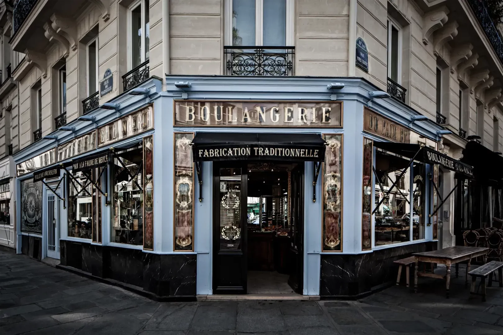 Paris'te köşe başında konumlanan, Du Pain et des Idées adını taşıyan, Fabrication Traditionnelle yazılı tabelasıyla geleneksel üretim vurgulayan, mavi detaylı ve süslü vitrinli boulangerie binasının dış cephesi, kaldırımdaki ahşap masa ve banklarla zarif bir görünüm sunmaktadır.
