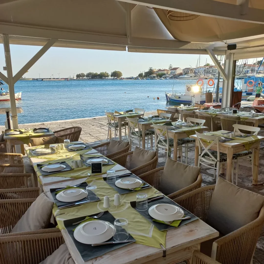 Deniz kenarında, açık hava bir restoranda, mavi denize karşı sıralanmış, yeşil ve sarı desenli masa düzenlemeleriyle donatılmış ahşap masalar ve rahat hasır sandalyeler, uzakta demirli tekneler ile bir sahil kasabasının binaları eşliğinde güneşli bir günde davetkar bir yemek ortamı sunuyor.