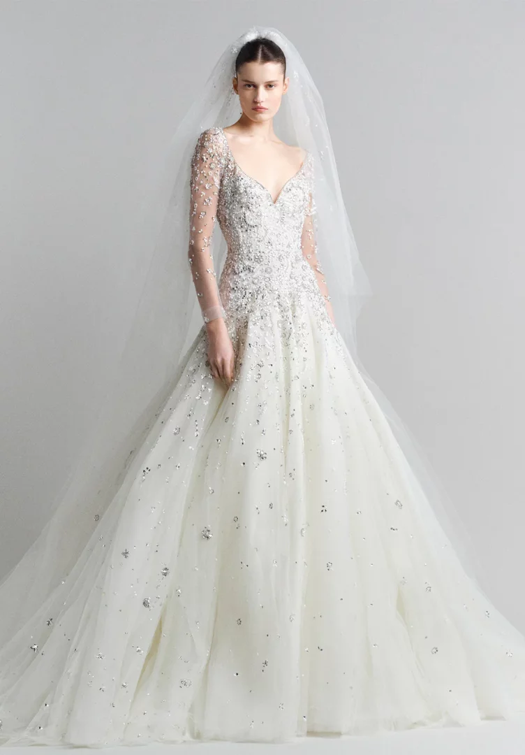 Elie Saab'ın Bahar 2027 gelin koleksiyonundan, bedeni ve uzun şeffaf kolları gümüş payet ve boncuklarla yoğun şekilde işli, kabarık eteği ise parıltılı detaylarla süslenmiş, zarif beyaz gelinliği giyen bir kadın model.