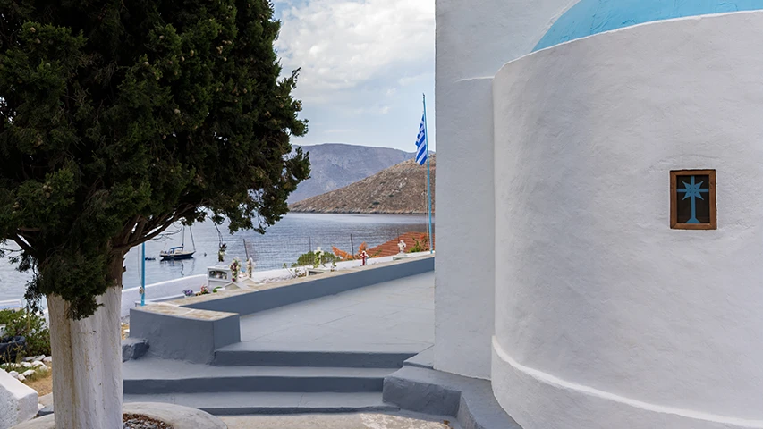 Kalymnos'un Emporios Koyu'nda, yeşil bir selvi ağacının önünden görünen, mavi kubbeli ve pencereli haç detaylı bembeyaz bir şapel ile körfezdeki teknelerin ve karşıdaki tepelik arazinin panoramik manzarası, hafif bulutlu gökyüzü altında huzurlu bir atmosfer sunuyor.