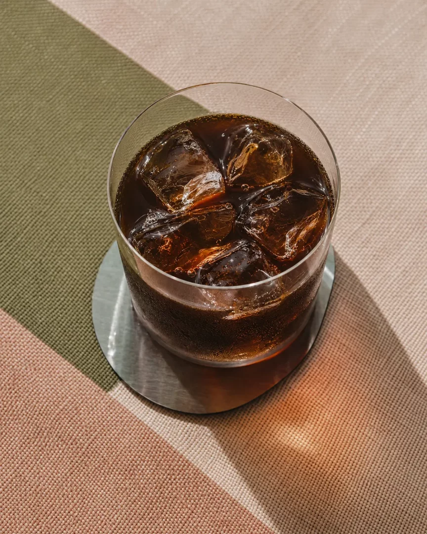 Soğuk ve ferahlatıcı bir görünüm sunan, bol buz küpleriyle dolu, koyu renkli bir cold brew kahve bardağı; metal bir altlık üzerinde dururken, zeytin yeşili, bej ve şeftali tonlarındaki kumaş zemine düşen güneş ışıkları ve keskin gölgeler, bu eşsiz cold brew tarifinin keyfini çıkarmaya davet ediyor.