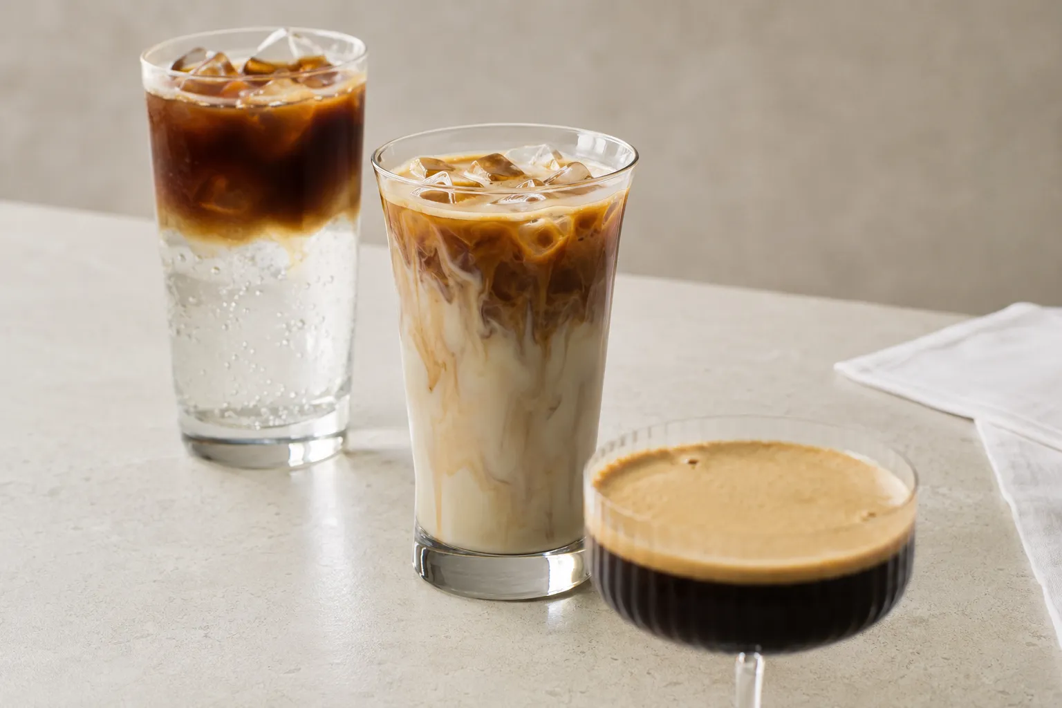 Açık renkli bir tezgah üzerinde, altta gazlı su üstte koyu espresso katmanıyla buzlu bir içecek, mermer deseni oluşturan espresso ve süt karışımıyla hazırlanan buzlu latte ile üzerinde kalın krema tabakası olan ayaklı bir kadehte sunulan yoğun bir soğuk espresso olmak üzere üç farklı espresso bazlı soğuk kahve sergileniyor.