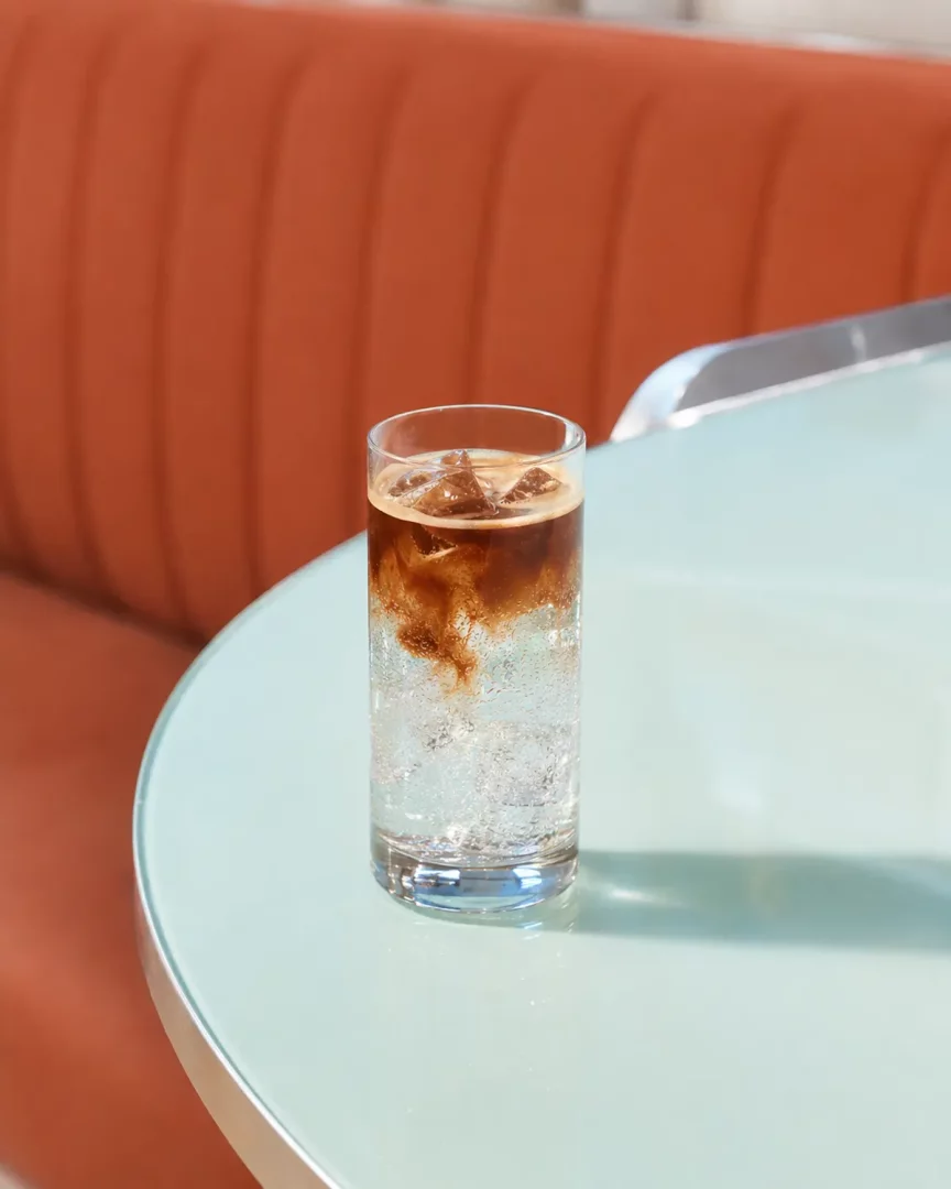 Açık mint yeşili bir masanın üzerinde, içinde bol buz ve gazlı su bulunan uzun bir bardağa yeni eklenmiş koyu renkli espresso'nun nazikçe karıştığı, adeta bulutlar oluşturan bir espresso tonic içeceği net bir şekilde görülüyor; arka planda sıcak turuncu tonlarında döşemeli bir koltuk var.