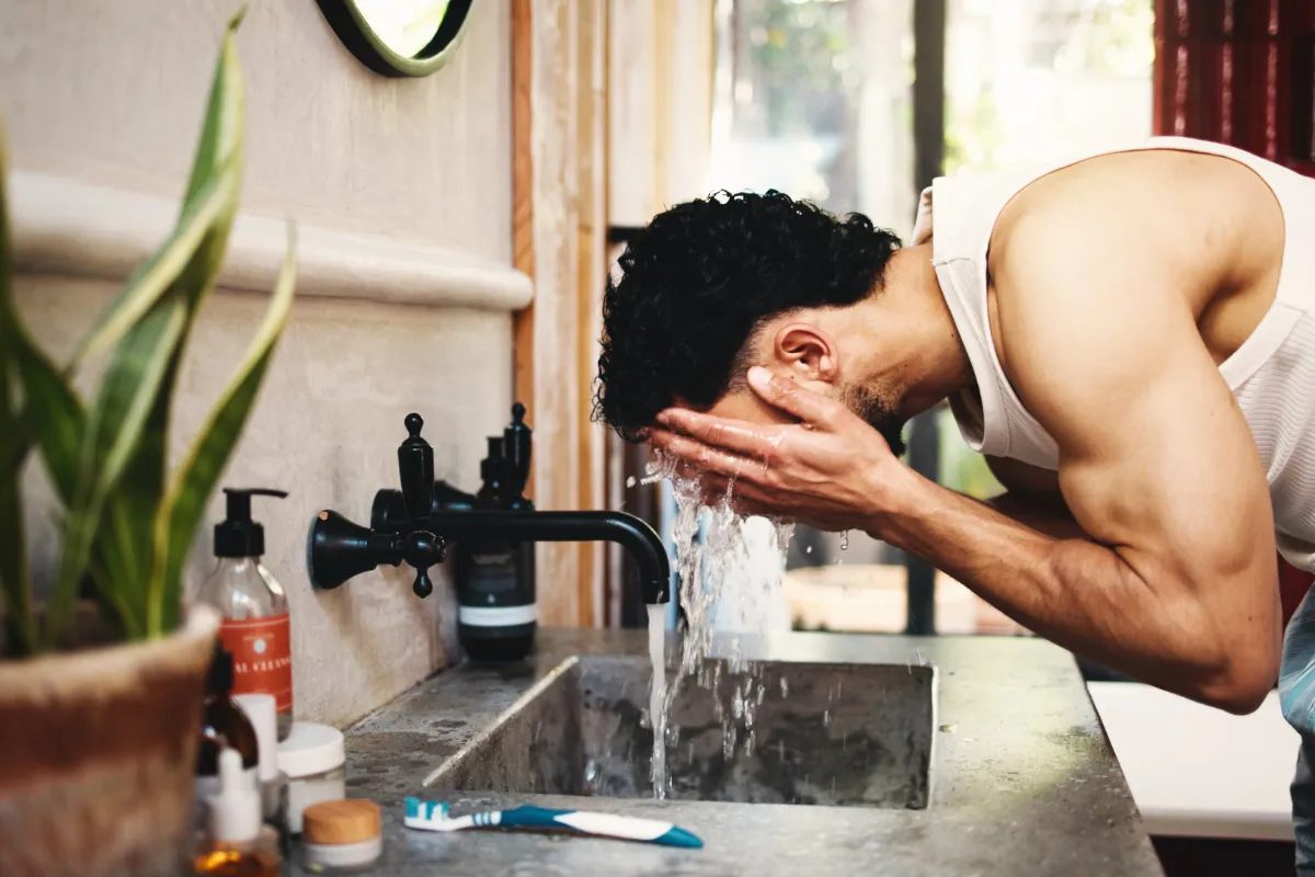 Kıvırcık saçlı, sakallı bir adam, beyaz atletiyle, modern banyo lavabosunda yüzünü yıkamak için musluktan akan suyu avuçlarında toplarken, etraftaki ürünler yüz temizleme rutinlerinin önemini hatırlatıyor.