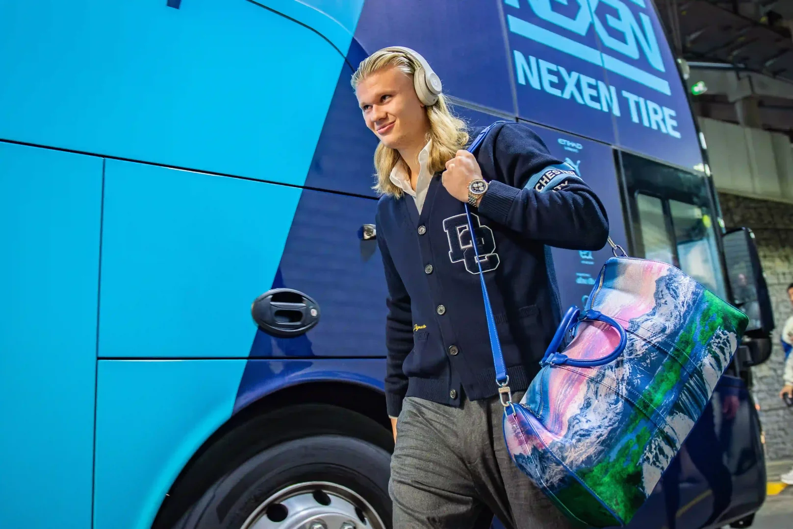 Erling Haaland, Manchester City'nin genç yıldızı, kulaklıkları takılı uzun sarı saçlarıyla, üzerinde büyük bir D harfi arması bulunan lacivert hırkası ve rengarenk dağ manzaralı omuz çantasıyla takım otobüsünün yanında gülümseyerek yürüyor; otobüsün üzerinde belirgin Nexen Tire ve Etihad Airways yazıları görülebiliyor.