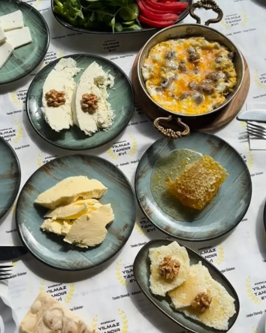 Erzincan'daki en iyi kahvaltı deneyimlerinden birini sunan Yılmaz Tandır Evi'nde, masayı dolduran petek balı, dilimlenmiş doğal tereyağı, demir tavada servis edilen kavurmalı yumurta, cevizli tulum peyniri, beyaz peynir ve ballı cevizli ekmek dilimleri gibi yöresel lezzetlerin bir araya geldiği zengin ve iştah açıcı bir Türk kahvaltısı sofrası.