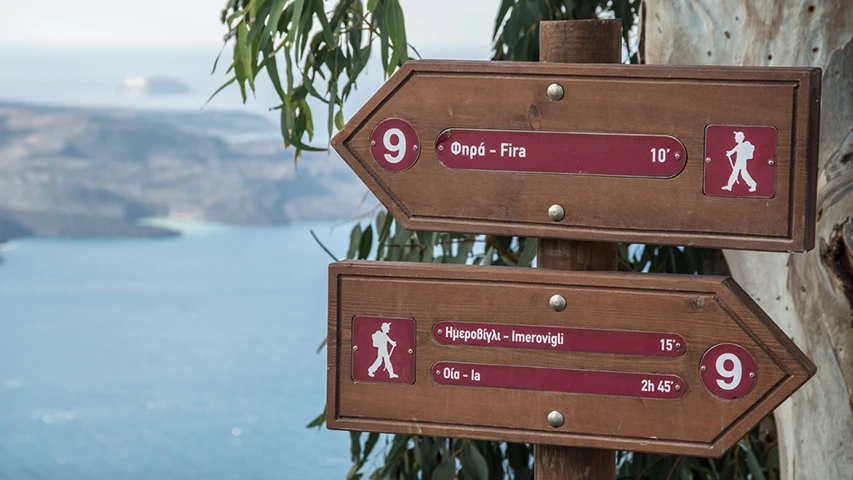 Fira-Oia yolu üzerinde, deniz manzaralı bir noktada konumlanan, ahşap bir direğe sabitlenmiş ok şeklindeki iki yön tabelası, Fira'ya 10 dakika ve Oia'ya 2 saat 45 dakika yürüyüş mesafelerini göstererek patika bilgilerini aktarıyor.
