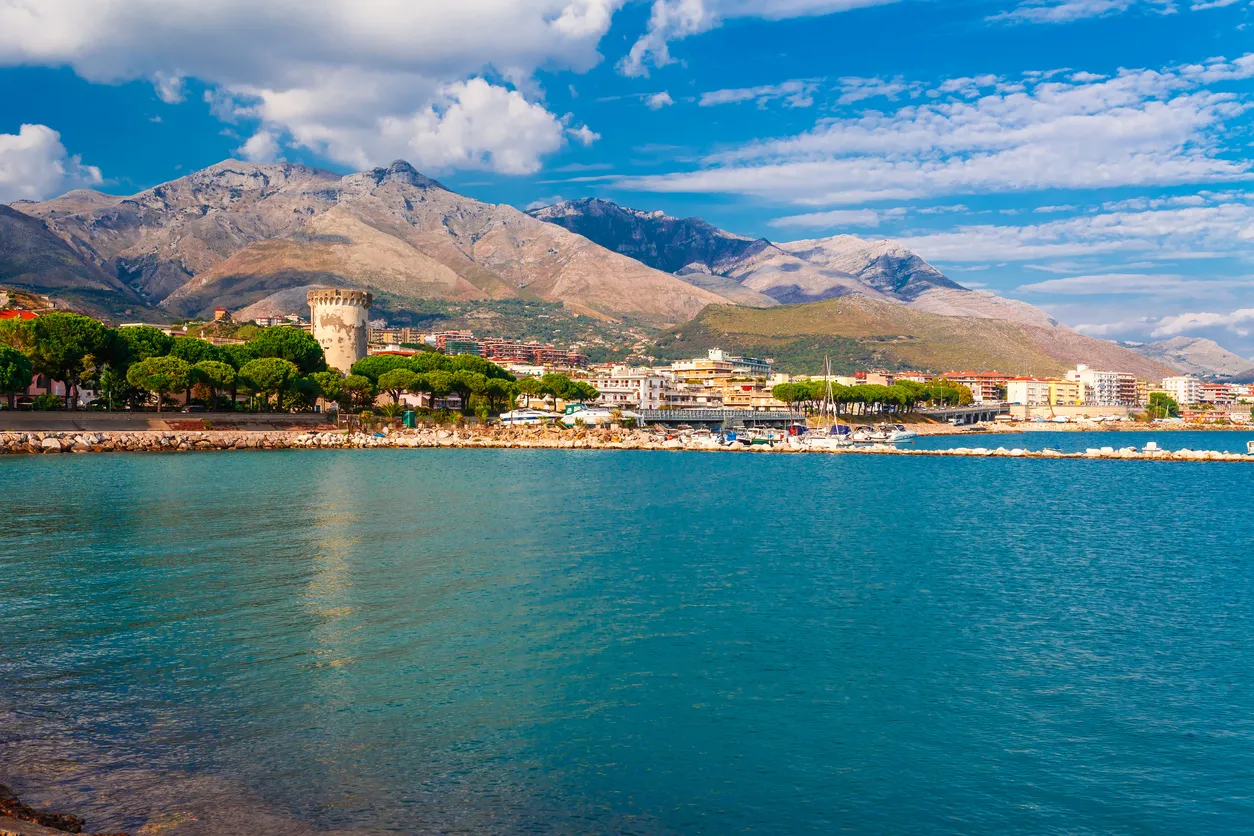 Parlak turkuaz deniz, kıyısında eski bir taş kulenin ve yemyeşil ağaçların sıralandığı Formia'nın hareketli sahil kasabasını gözler önüne sererken, arka planda görkemli dağlar ve parçalı bulutlu mavi gökyüzü yükseliyor.