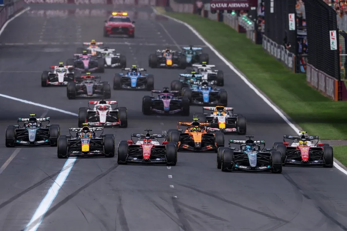 Beyaz çizgili bir yarış pistinde yan yana sıralanmış, farklı takımlara ait kırmızı, beyaz, mavi ve turuncu gibi çeşitli renklerde çok sayıda Formula 1 aracı, büyük bir heyecanla yarışa başlamak için hazır bekliyor, bu sahne Formula 1'in takvimindeki bir yarışı temsil ediyor.