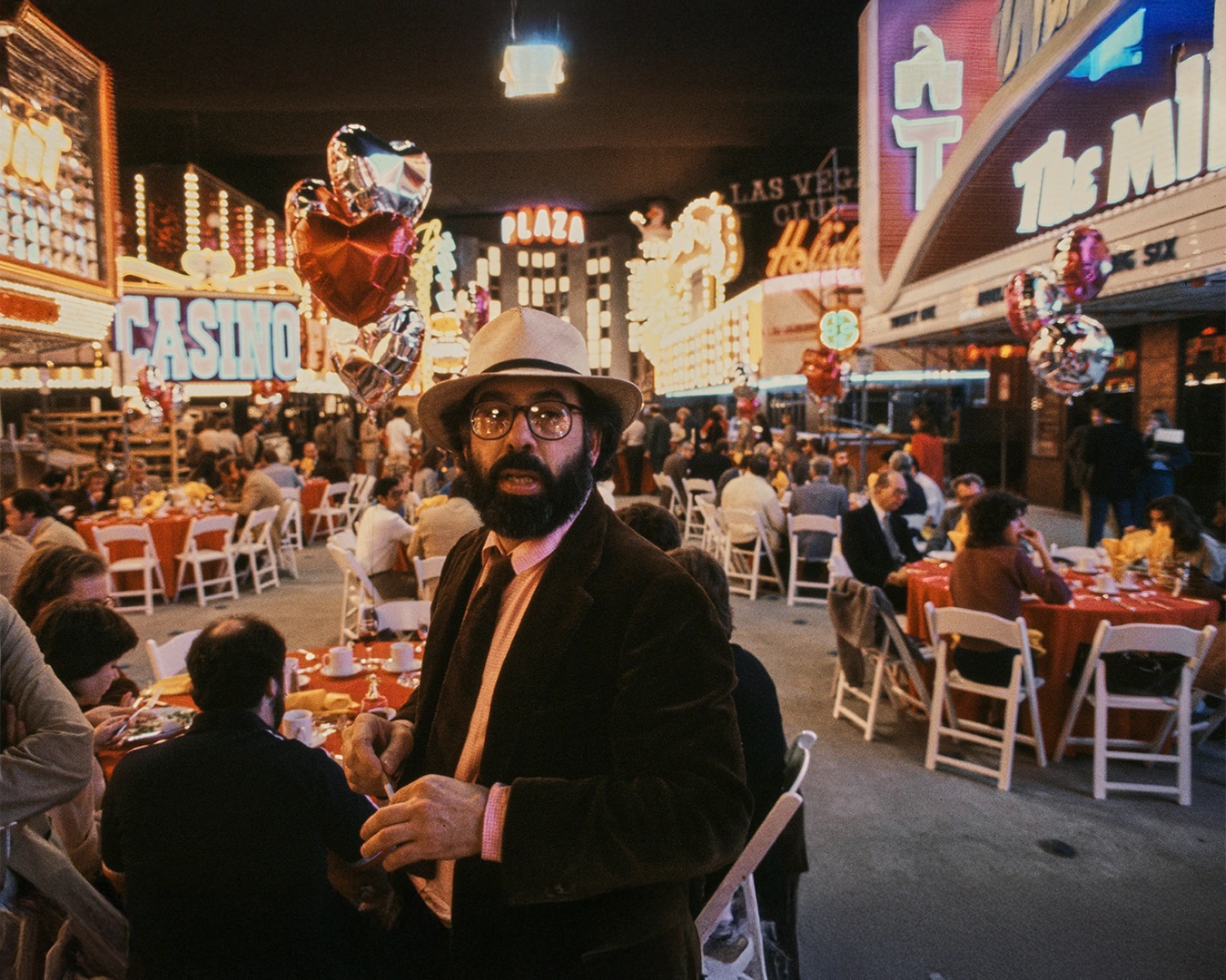 Şapkalı, sakallı ve gözlüklü usta sinema yönetmeni Francis Ford Coppola, Las Vegas'ın ışıltılı atmosferinde, neon ışıklı kumarhane ve otel tabelaları ile kalabalık masalar arasında, direkt olarak kameraya bakarken görüntüleniyor.