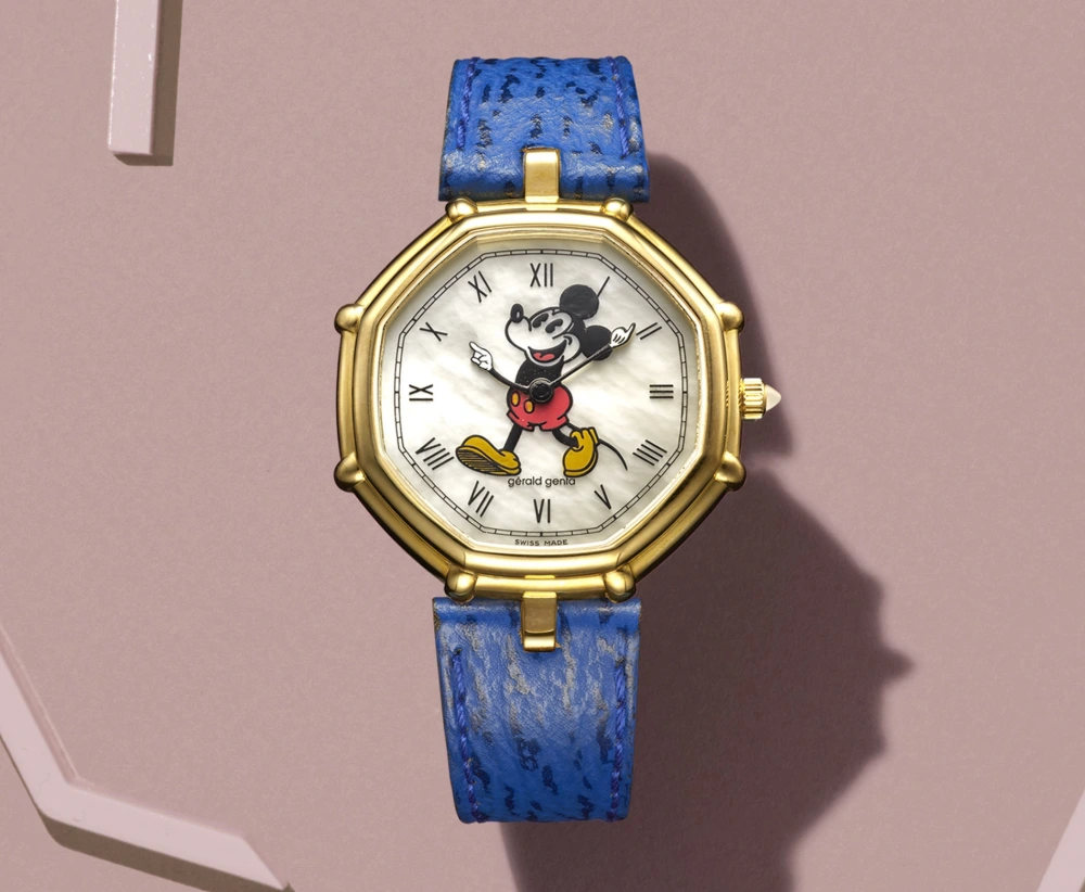 Açık morumsu pembe bir fonda sergilenen, altın rengi sekizgen kasalı Gerald Genta kol saati; sedef kadranında Romen rakamları ve kolları akrep yelkovan görevi üstlenen canlı renklerde Mickey Mouse figürüyle, mavi dokulu kayışıyla öne çıkıyor.