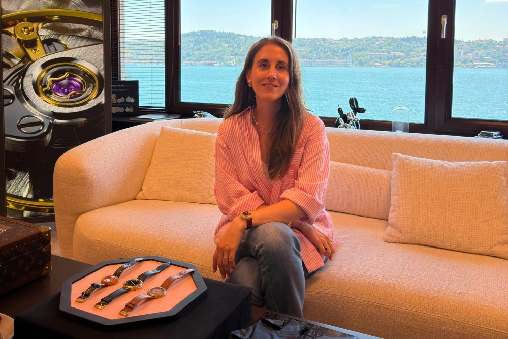 Lüks saatçilik dünyasının önemli isimlerinden Gerald Genta'nın kızı Alexia Genta, Boğaz manzaralı lüks bir ofiste açık renkli bir koltukta otururken, önündeki sehpa üzerinde sergilenen deri kayışlı birkaç lüks saatle birlikte gülümseyerek objektife bakıyor.
