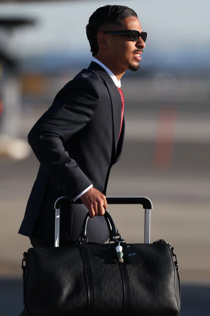 Fas takımının oyuncusu Gessime Yassine, şık koyu takım elbisesi, kırmızı kravatı, güneş gözlüğü ve bakımlı saç-sakal tarzıyla gülümseyerek, Louis Vuitton desenli siyah deri seyahat çantasını ve tekerlekli valizini taşırken, Dünya Kupası yolculuğu için uçağa doğru yürüyor.