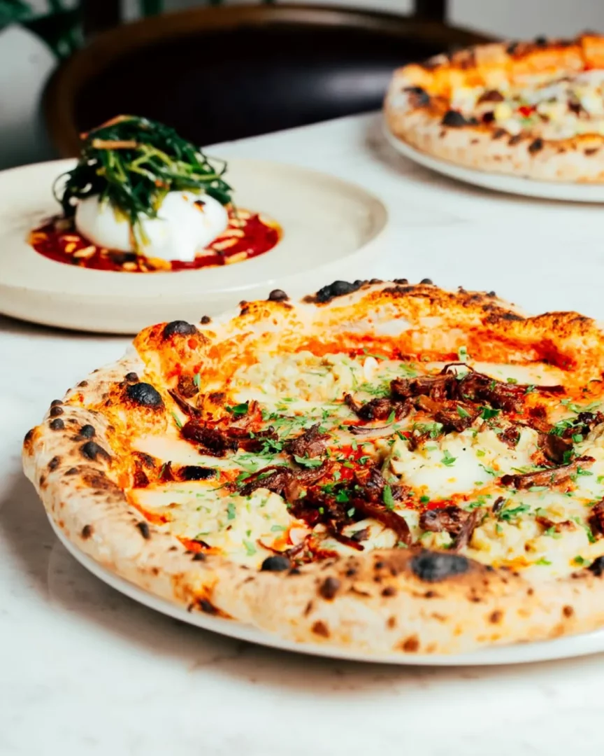 Yeldeğirmeni'ndeki bir restoranda, hafif yanık ve kabarık kenarlı, üzerinde beyaz peynir, karamelize soğan ve taze otlar bulunan lezzetli bir pizzanın ön planda olduğu; arkada burrata peyniri, çam fıstığı ve yeşilliklerle sunulan başka bir mezenin ve bulanık bir pizzanın daha göründüğü iştah açıcı bir masa düzeni.