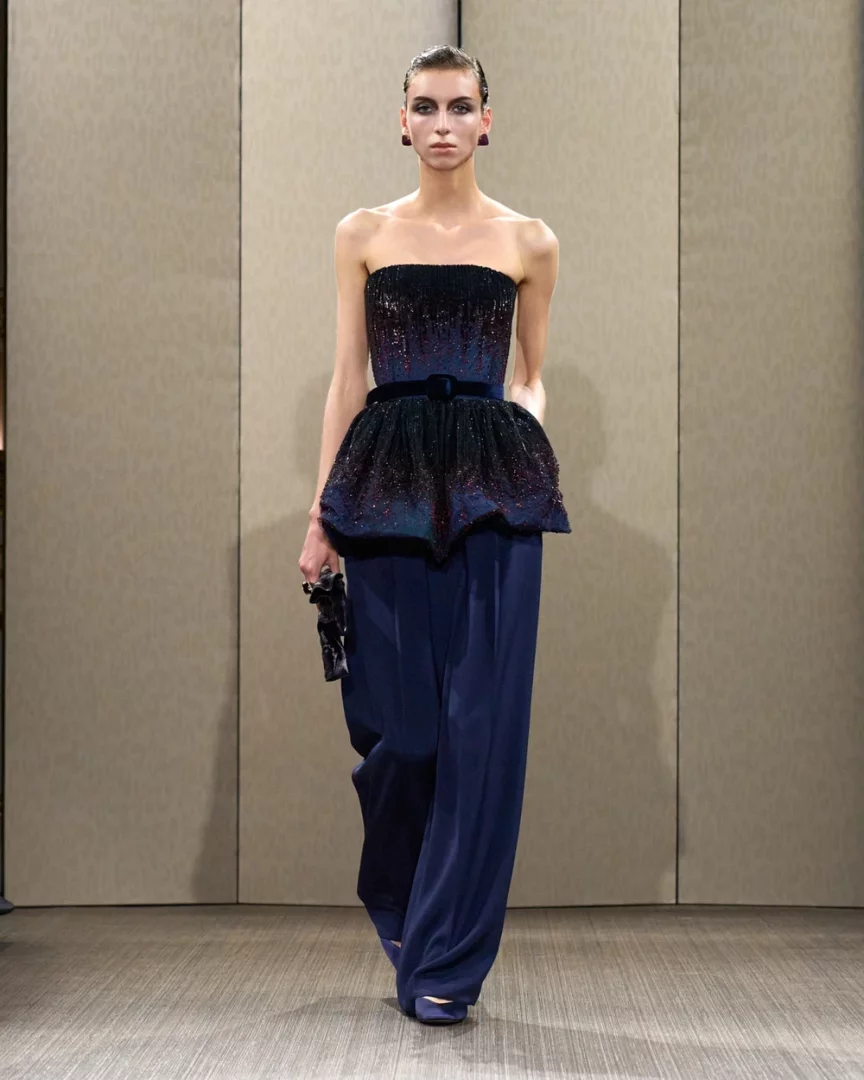 Paris Haute Couture Moda Haftası Sonbahar-Kış 2026 defilesinden bir karede, bir model podyumda Giorgio Armani Privé imzalı, lacivert tonlarında, ışıltılı ve taş işlemeli straplez peplum bluz ile bol paça pantolondan oluşan göz alıcı bir couture tasarımı sergiliyor.