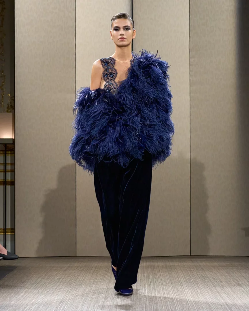 Paris Haute Couture Moda Haftası'nda Giorgio Armani Privé'nin Sonbahar-Kış 2026 koleksiyonundan, tek omzu zarif işlemelerle süslü, diğer omzu ve kolu kabarık lacivert tüylerle bezeli bir bluz ve kadife geniş paça pantolondan oluşan iddialı bir kıyafetle podyumda yürüyen model.