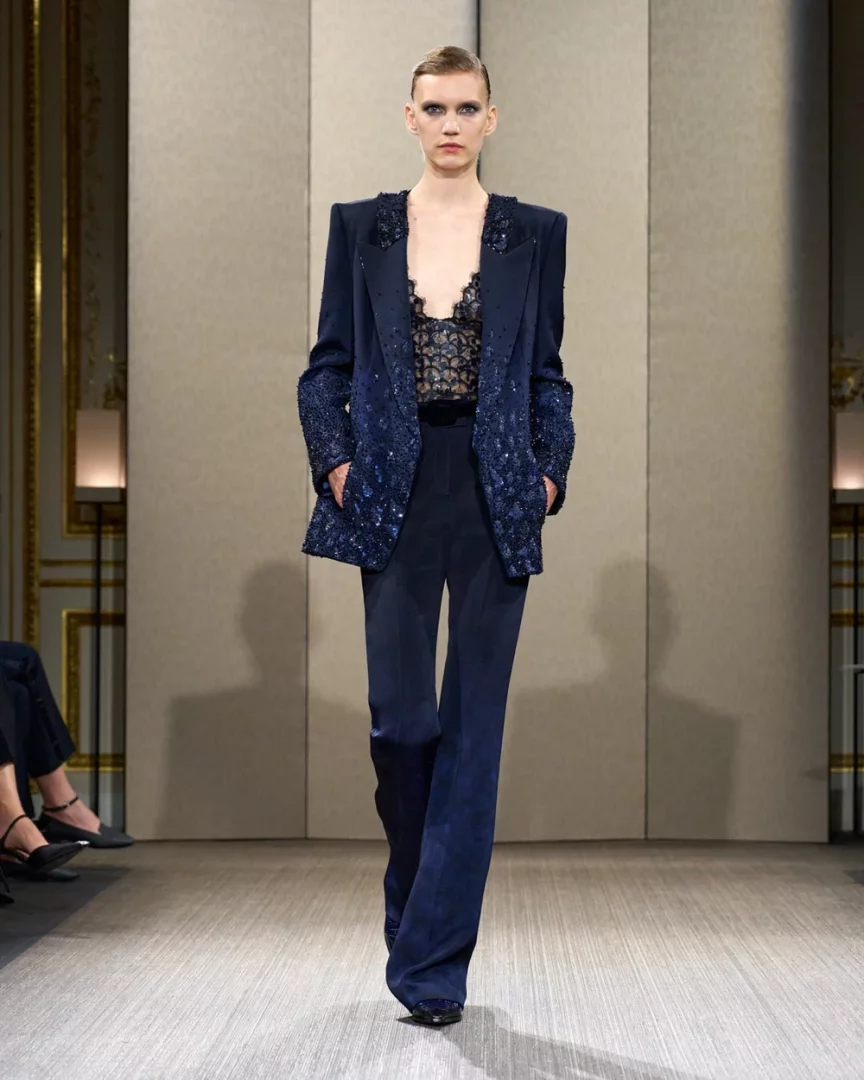 Paris Haute Couture Moda Haftası Sonbahar-Kış 2026 kapsamında Giorgio Armani Privé defilesinden bir görünüm sunan bu karede, omuzları belirgin, alt kısmı ve kolları yoğun pul ve işleme detaylarıyla süslü koyu lacivert ceket ile İspanyol paça pantolon giyen bir model podyumda yürüyor, ceketinin içinde dantel büstiyer dikkat çekiyor.
