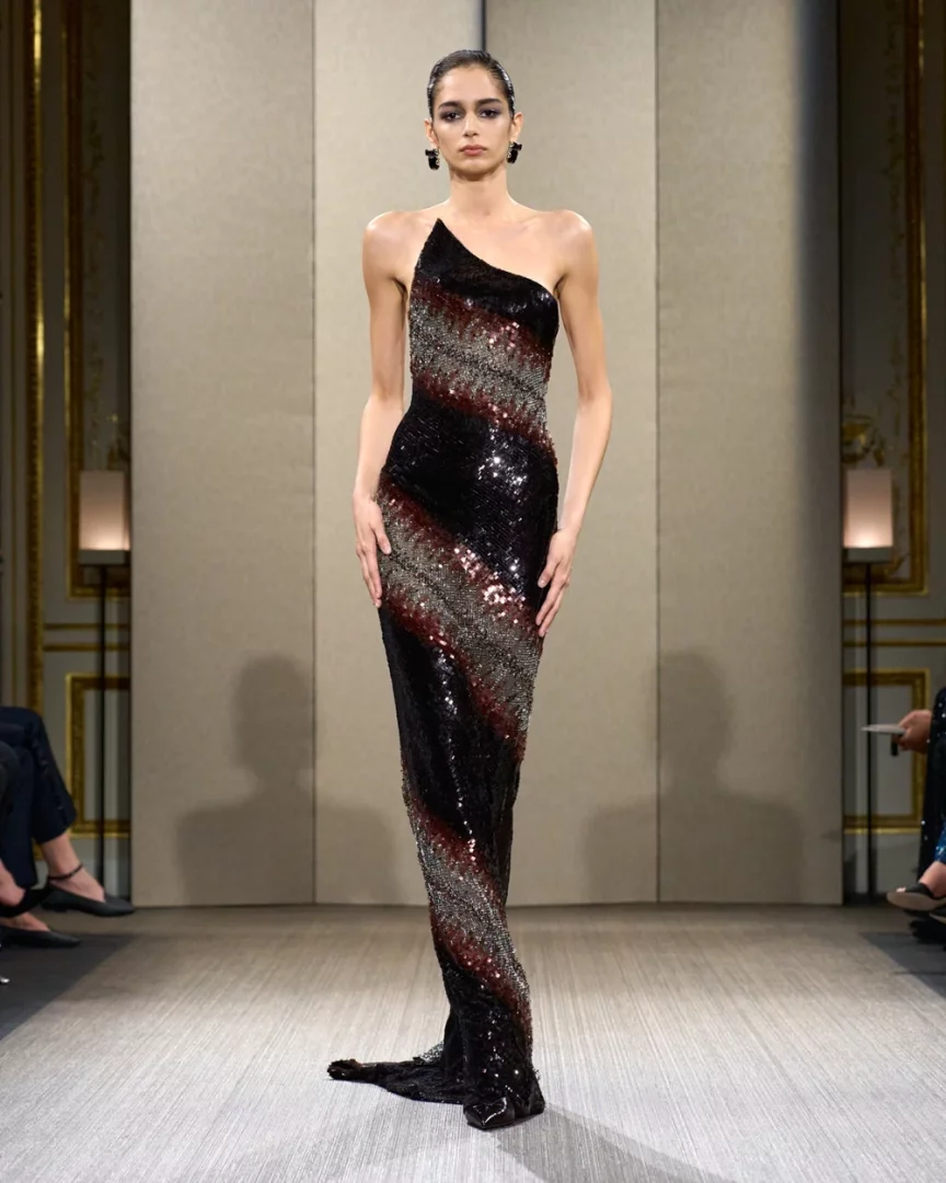 Bir model, Paris Haute Couture Moda Haftası'nda sergilenen Giorgio Armani Privé Sonbahar-Kış 2026 couture koleksiyonundan, siyah, koyu kırmızı ve gümüş payetlerle oluşturulmuş dinamik çapraz çizgilere sahip, tek omuzlu, yere kadar uzanan, podyumda göz kamaştıran bir gece elbisesiyle poz veriyor.