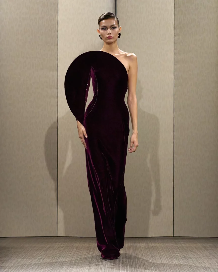 Paris Haute Couture Moda Haftası Sonbahar-Kış 2026 kapsamında Giorgio Armani Privé koleksiyonundan, podyumda zarifçe duran bir model; tek omuzlu, sol omzunda mimari bir daire formunda dramatik ve heykelsi bir detay barındıran, koyu bordo kadife uzun bir haute couture elbise sergiliyor.