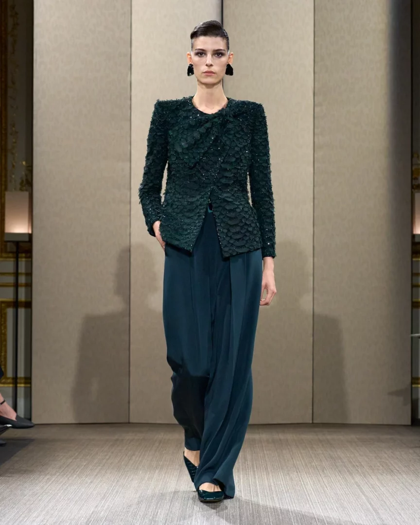 Bir model, Paris Haute Couture Moda Haftası'nda Giorgio Armani Privé Sonbahar-Kış 2026 couture koleksiyonundan, pul işlemeli koyu yeşil bir ceket ile akışkan bol paça pantolondan oluşan sofistike bir takımı podyumda sunarken, keskin omuz hatları ve dramatik küpeleriyle dikkat çekiyor.