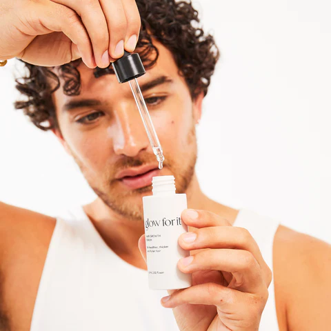 Kıvırcık saçlı genç bir erkek, elinde glow for it markalı saç büyüme serumu şişesini dikkatle tutuyor ve damlalık aracılığıyla şişeye tek bir damla damlatmaya hazırlanırken, ürünün daha sağlıklı, kalın ve dolgun saçlar için olduğunu gösteren etiketi belirgin bir şekilde görülüyor.