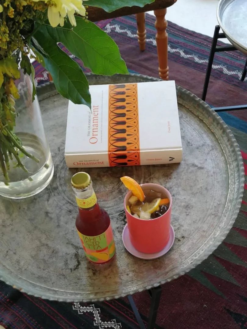 Eskitme görünümlü metal bir tepsi üzerinde, Tahiti buzlu çay, meyve dilimli serinletici bir içecek ve The Grammar of Ornament adlı kitabın bulunduğu bu detay, Göcek'te geçirilen keyifli ve dinlendirici bir tatil anını akla getiriyor.