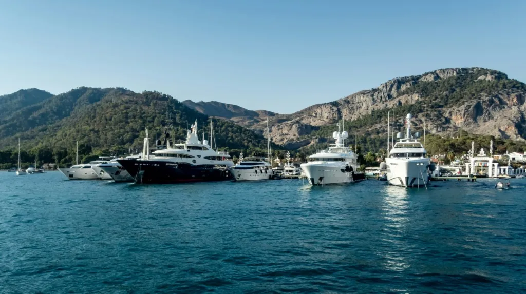 Göcek koyunda, masmavi berrak sularda demirlemiş, büyüleyici yeşil dağlarla çevrili birçok lüks yatın ve teknenin huzur dolu görüntüsü, Göcek'in en popüler gezilecek yerlerinden birinin keyifli atmosferini yansıtıyor.