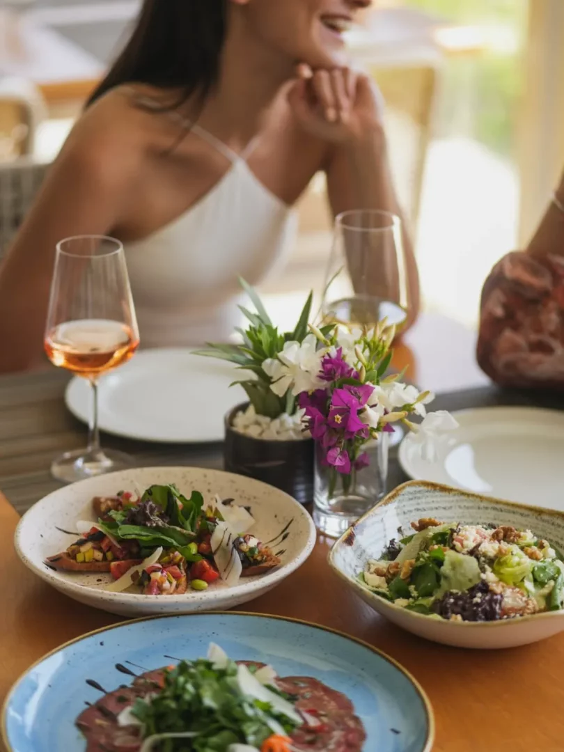 Göcek gezilecek yerler arasında yer alan şık bir restoranda, masada roka ve parmesanlı carpaccio, cevizli salata ve bruschetta benzeri çeşitli başlangıçlar ile pembe şarap eşliğinde keyifli bir öğün anında gülümseyen bir kadın görülüyor.