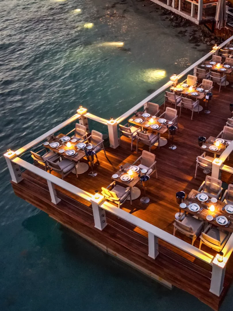 Göcek gezilecek yerler arasında romantik bir akşam yemeği için ideal, denizin üzerinde yer alan ahşap iskeledeki şık bir açık hava restoranı; masalar mum ışıklarıyla aydınlatılmış, etraftaki loş ambiyans ve suyun üzerindeki yansımalar huzurlu bir atmosfer sunuyor.