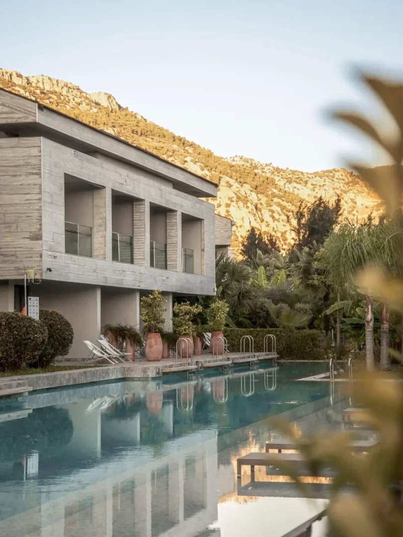Günbatımı ışıklarıyla aydınlanan kayalık dağların eteklerinde yer alan, modern mimariye sahip bir otel veya tesisin balkonlu yapısı ve önündeki berrak yüzme havuzunun yansımaları, Göcek'in doğal güzelliklerini ve konaklama seçeneklerini vurguluyor.